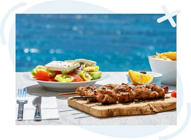 Greek meal by the sea with skewered grilled meat on a wooden board, a village salad with feta, and fries in the background.