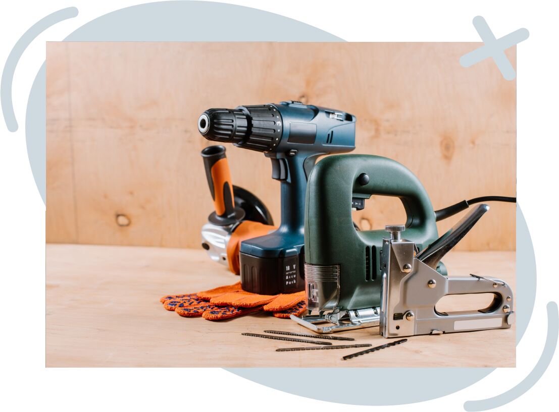 Assortment of power tools on a wooden workbench, including a cordless drill, jigsaw, angle grinder, and staple gun with blades and safety gloves.