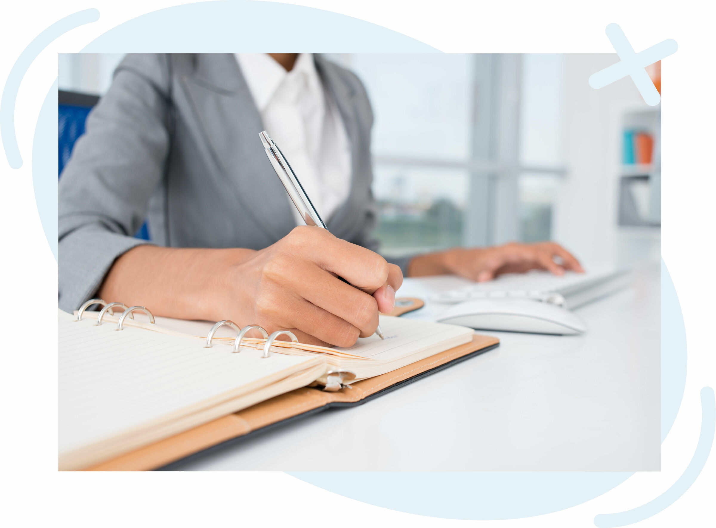 Person in a gray blazer writing in a ring-bound notebook at a desk while using a computer mouse and keyboard.