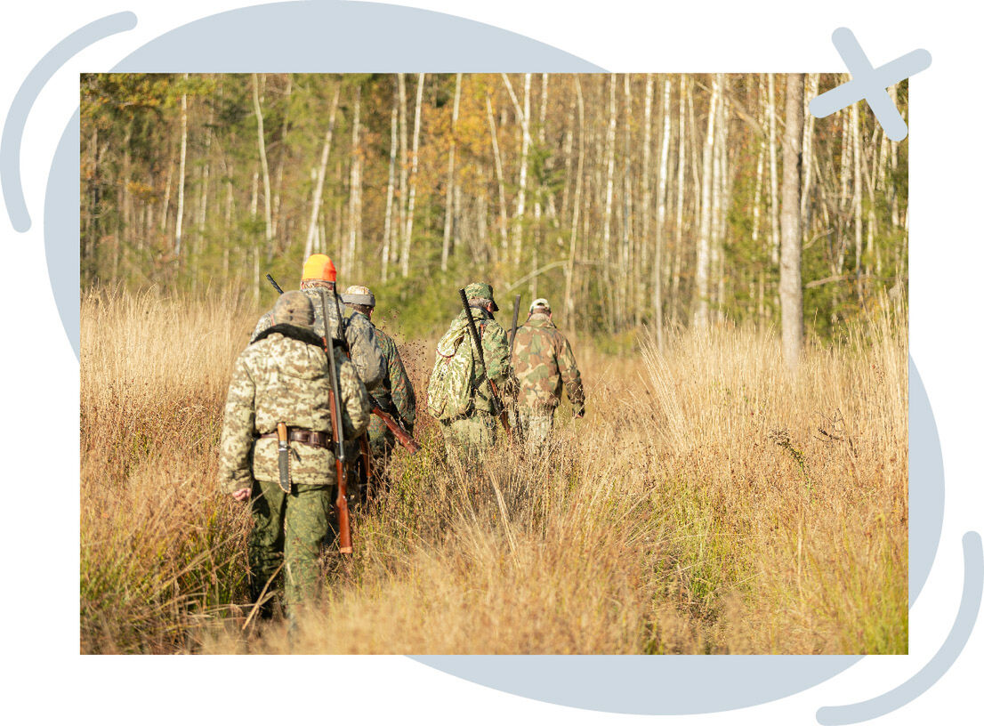 Group of hunters in camouflage walking single file through tall dry grass toward a forest, rifles slung over their shoulders.