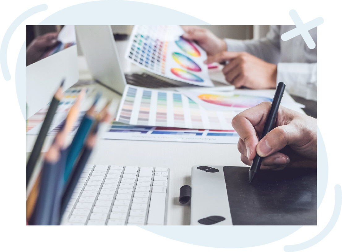 Close-up of a design workspace with a person drawing on a graphics tablet beside a keyboard, while another hand holds color swatches and a color wheel chart near a laptop.