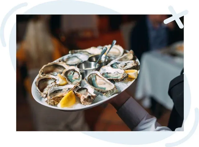 Server holding a round platter of fresh oysters on crushed ice with lemon wedges and a metal sauce cup, set in a softly lit restaurant.