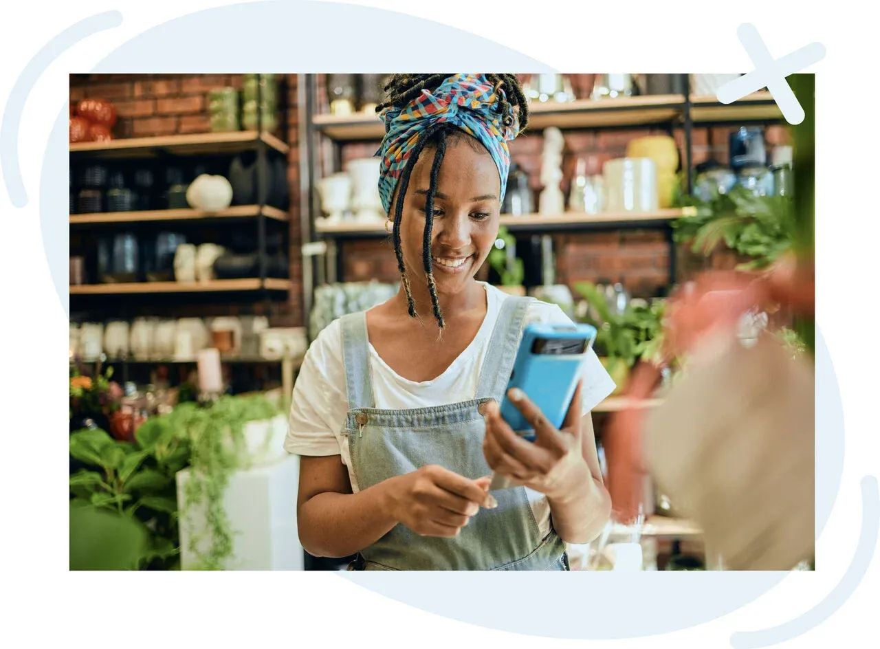 Smiling shop worker in a plant store looking at a smartphone while standing among greenery and shelves of pots.
