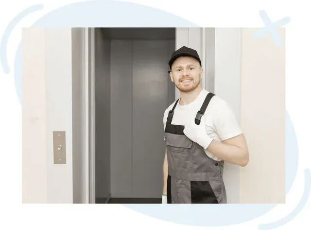 Smiling maintenance worker standing beside an open elevator doorway, wearing overalls, a white t‑shirt, gloves, and a black cap.
