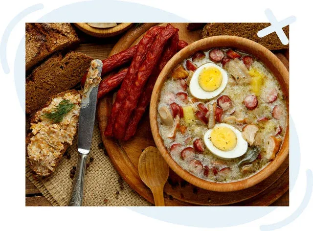 Rustic Polish meal with sour rye soup topped with boiled eggs and sausage, served alongside smoked sausages, rye bread, and a spread on a wooden board.