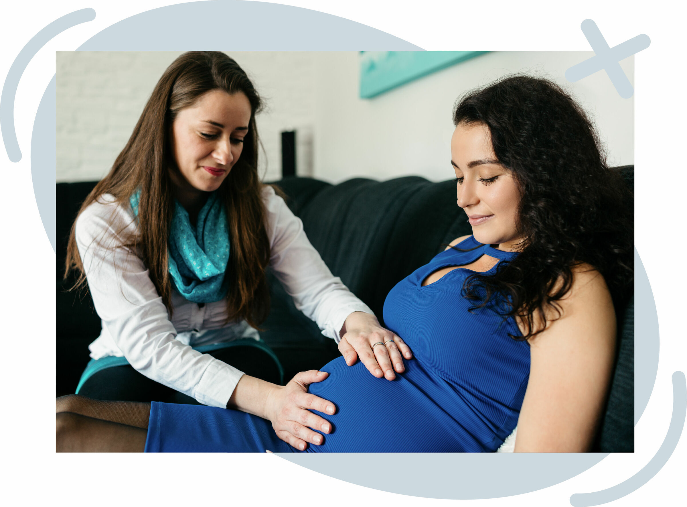Pregnant woman in a blue dress sits on a couch while another woman gently places her hands on the baby bump, offering support and care.
