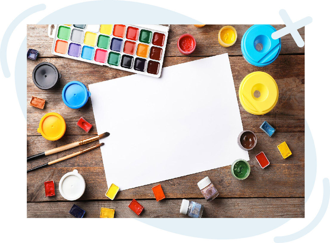 Top-down view of an art workspace with a blank white sheet of paper surrounded by watercolor paints, brushes, and small paint jars on a rustic wooden table.