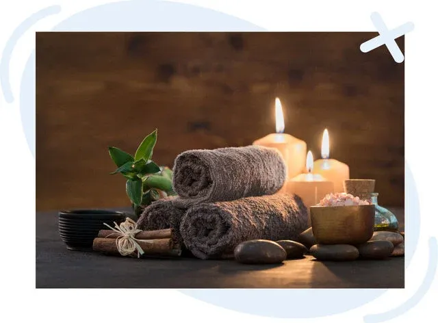 Cozy spa scene with rolled brown towels, lit candles, smooth stones, and bowls of bath salts on a dark surface.