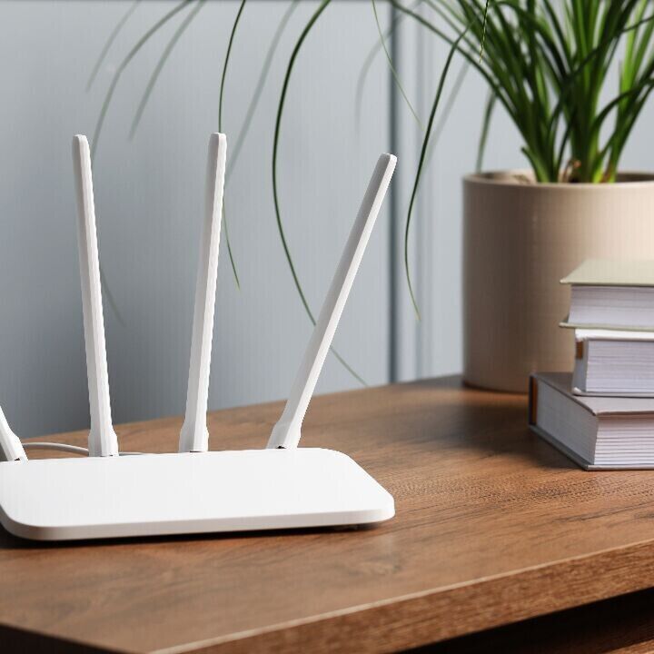 White Wi‑Fi router with four antennas on a wooden desk beside a stack of books and a potted plant.