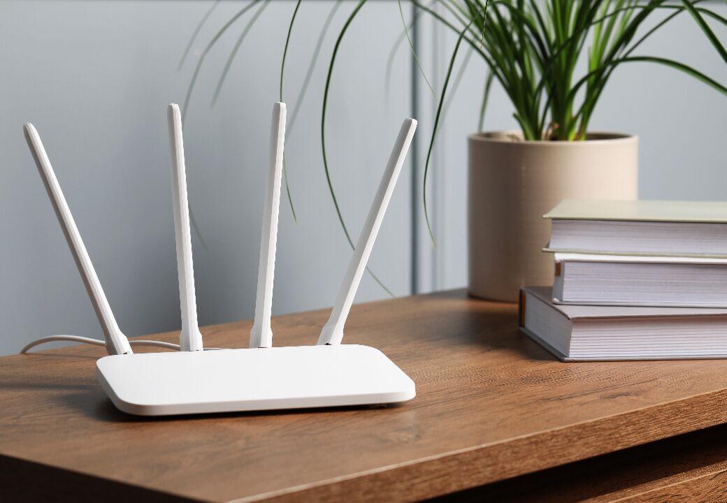 White Wi‑Fi router with four antennas on a wooden desk beside a stack of books and a potted plant.