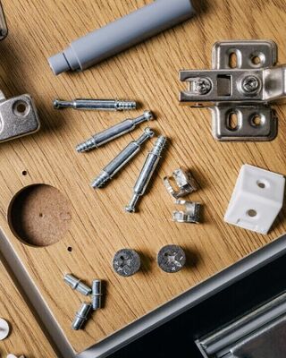Assorted cabinet hardware and fasteners laid out on wooden panels, including metal hinges, drawer slides, cam locks, screws, dowels, and plastic pieces.