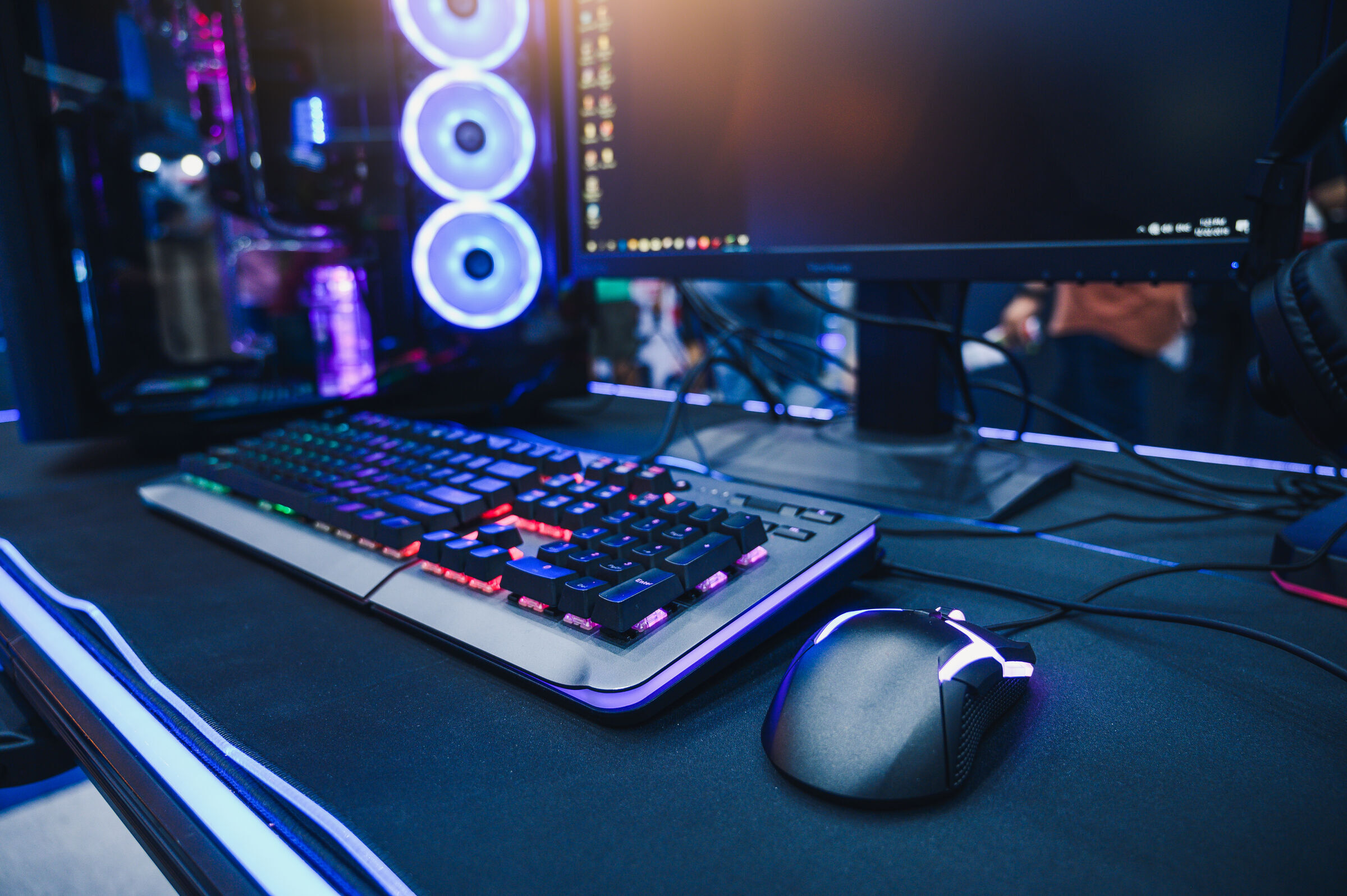RGB-lit gaming keyboard and mouse on a desk beside a monitor and PC with illuminated fans.