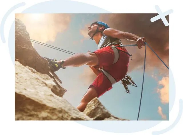 Low-angle view of a climber scaling a rocky cliff with ropes under a dramatic sky.
