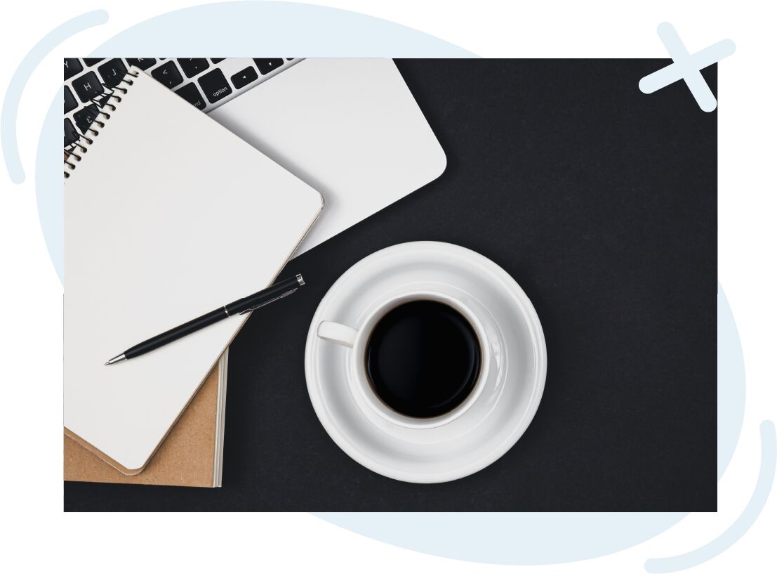 Top-down view of a workspace with a cup of black coffee, a notebook with a pen, and part of a laptop on a dark desk.
