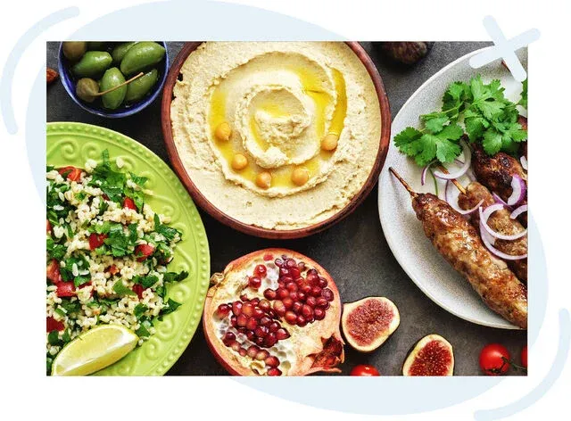 Assorted Middle Eastern dishes including a bowl of hummus drizzled with olive oil, tabbouleh salad, olives, grilled kofta skewers with onions and cilantro, and fruits like pomegranate and figs.
