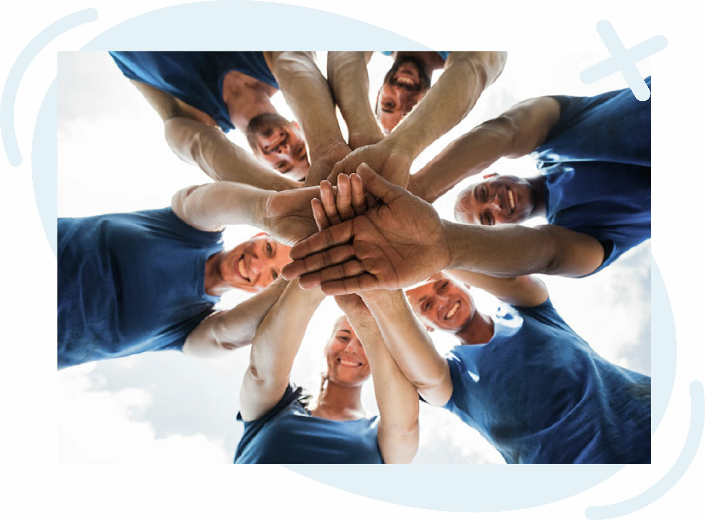 Group of people in blue shirts standing in a circle with hands stacked together, smiling and looking down toward the camera from a low-angle view against a bright sky.