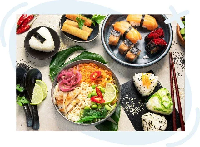 Assorted Japanese-inspired dishes on a tabletop, including sushi, onigiri, spring rolls, and a vibrant noodle bowl with vegetables and lime wedges.