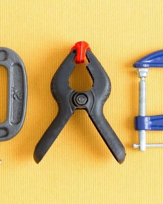 Three clamps arranged on a yellow textured surface: a gray C-clamp on the left, a black spring clamp with a red tip in the center, and a blue C-clamp on the right.