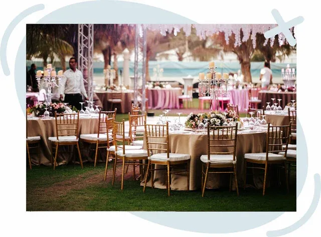 Elegant outdoor event with round tables draped in beige linens, gold chiavari chairs, floral centerpieces, and candelabras set on a grassy venue.