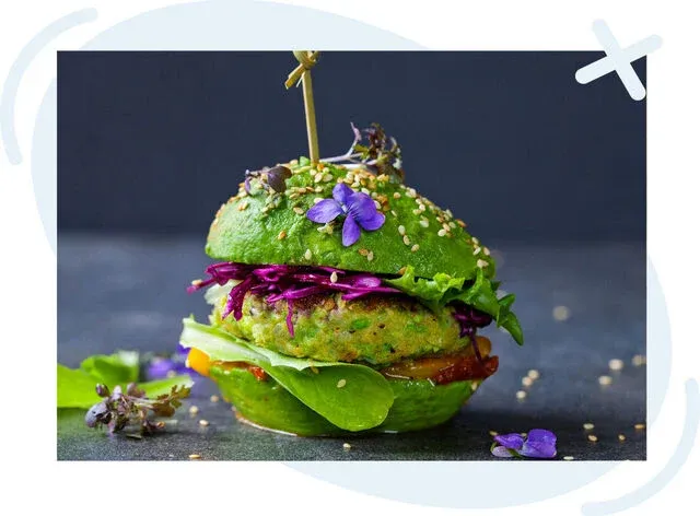 Colorful vegan burger with green bun, topped with sesame seeds and edible flowers, filled with greens and purple slaw on a dark background.