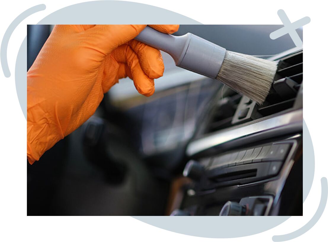 Gloved hand using a soft detailing brush to clean dust from a car’s air vent on the dashboard.