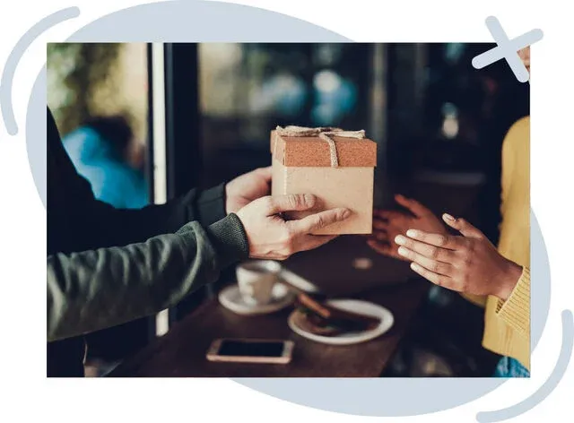 Close-up of two people exchanging a wrapped gift box over a cafe table, with coffee cups and a phone in the background.