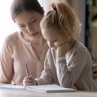 Adult helping a young girl with homework at a table, both focused on writing in a notebook.