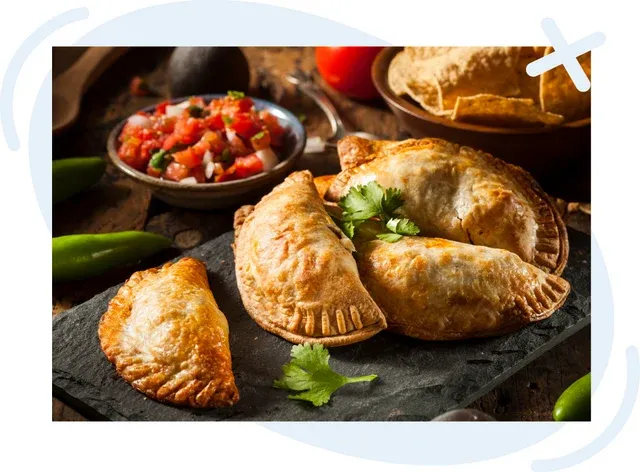 Rustic food scene with golden-brown empanadas on a dark slate board, garnished with cilantro, with a bowl of fresh salsa and other ingredients in the background.