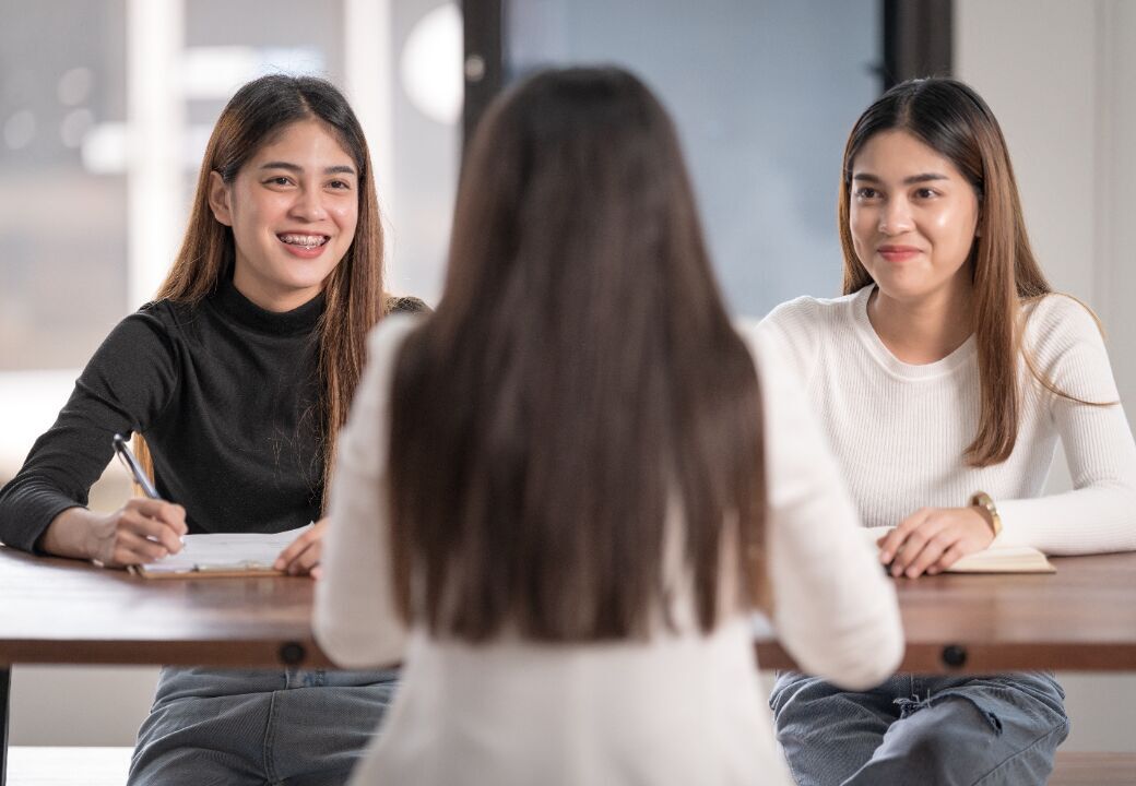 Two interviewers smile and take notes while speaking with a candidate seated across from them at a table.