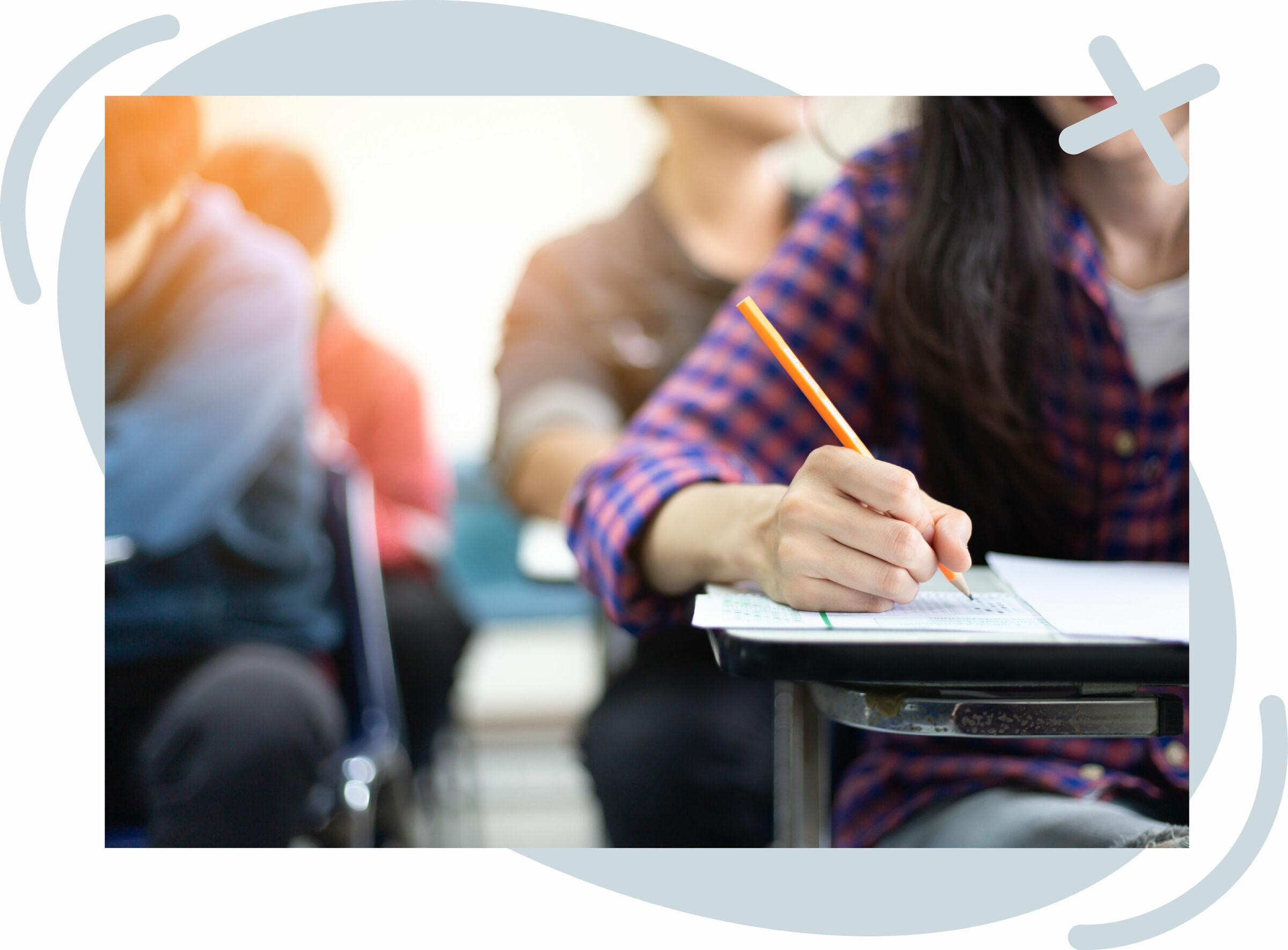 Close-up of a student filling out a test sheet with a pencil at a classroom desk, surrounded by other blurred students.