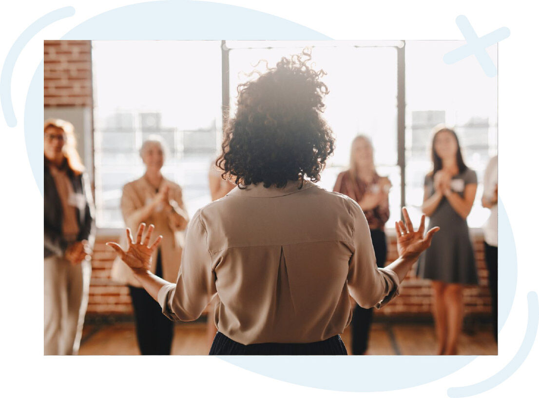 Coach speaking to a small group in a sunlit room while attendees stand in a semicircle and applaud.