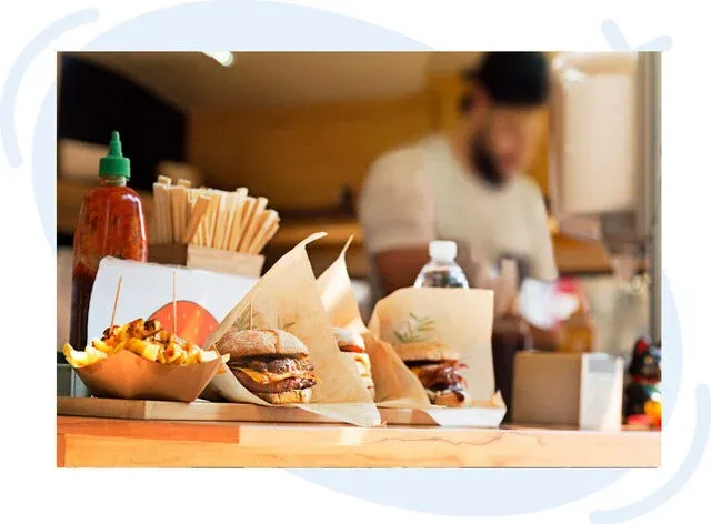 Close-up of a food truck counter displaying burgers, fries, and condiments with a blurred vendor in the background.
