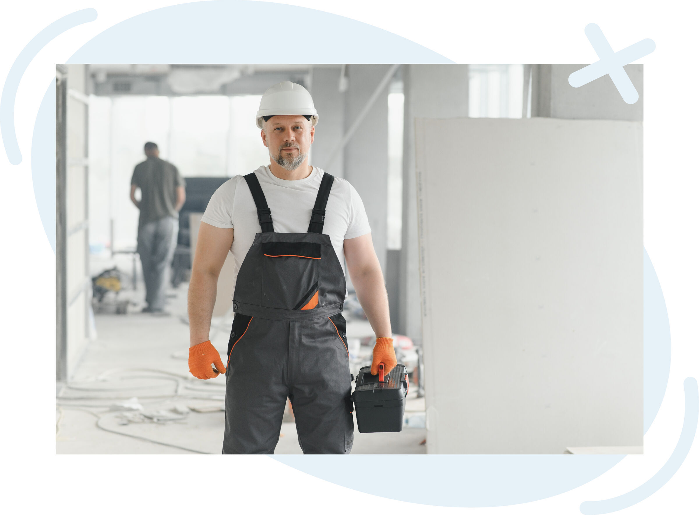 Construction worker in a hard hat and overalls holding a toolbox inside a bright, unfinished building.