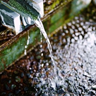 Close-up of water flowing from a metal spout into a shallow stone basin with pebbles, moss, and ripples.