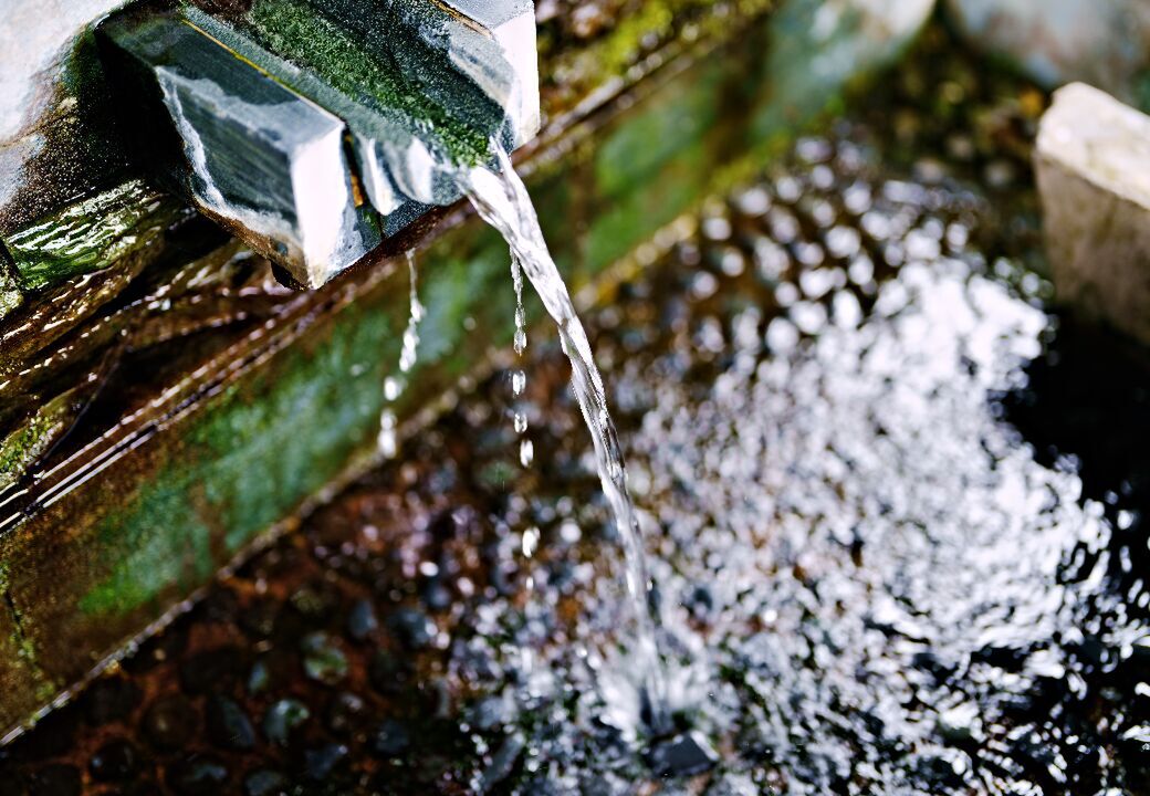 Close-up of water flowing from a metal spout into a shallow stone basin with pebbles, moss, and ripples.