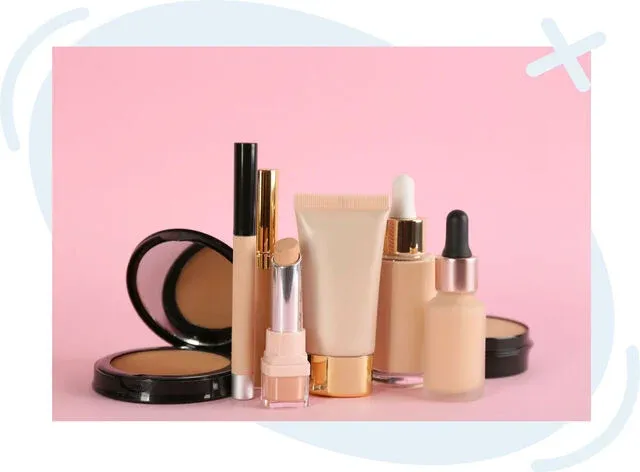Assorted nude-toned makeup products arranged on a pink background, including compact powders, a lipstick, concealer, a squeeze tube, and dropper bottles.