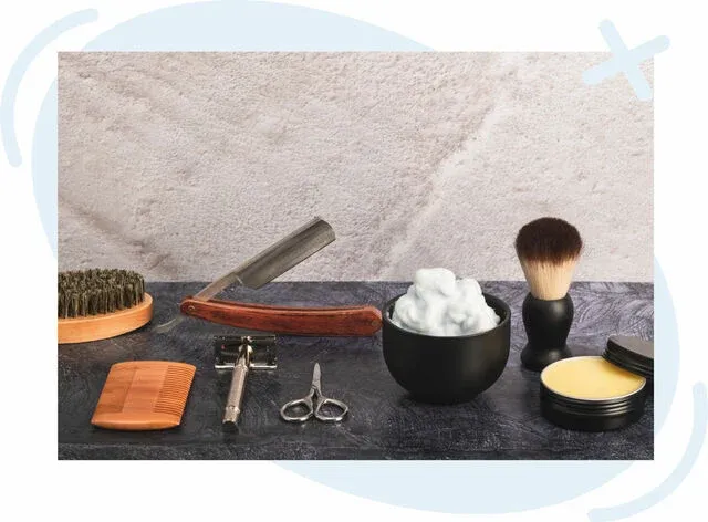 Assortment of traditional men’s grooming tools arranged on a dark countertop, including a shaving brush, safety razor, straight razor, comb, scissors, beard brush, bowl of shaving foam, and tin of balm.