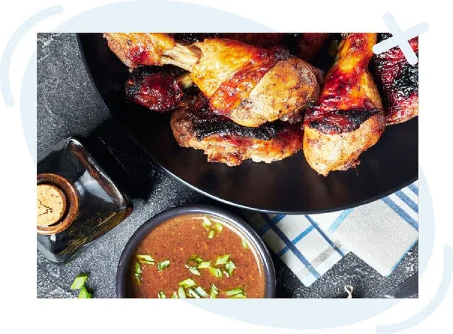 Close-up of grilled chicken drumsticks on a black plate with a bowl of brown dipping sauce and chopped herbs nearby.