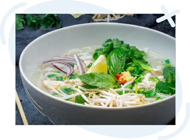Close-up of a steaming bowl of Vietnamese chicken pho garnished with fresh herbs, bean sprouts, red onion, chilies, and a wedge of lemon in a light, clear broth.