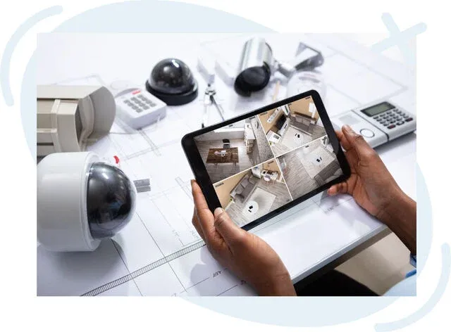Person monitoring a home security system on a tablet surrounded by various security cameras and devices on a desk with building plans.