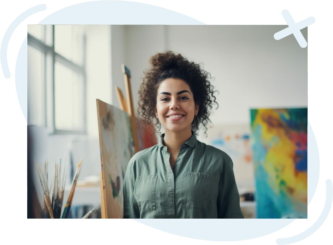 Smiling person standing in a bright art studio with canvases and paintbrushes around.