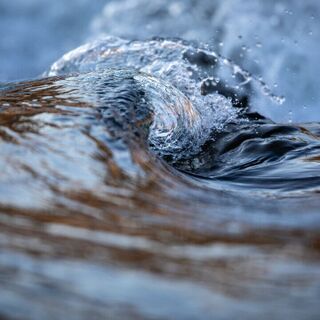 Close-up view of a small wave curling over in a stream, with crisp details of the water’s surface and splashes, rendered in cool blue and gray tones.