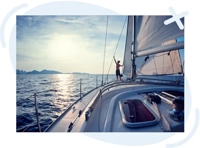 Person adjusting sails on a yacht at sea during a calm, bright evening with distant islands on the horizon.