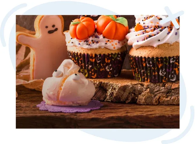 Assorted Halloween-themed treats including cupcakes topped with pumpkins and sprinkles, a ghost-shaped cookie, and a small ghost meringue on a rustic wooden surface.