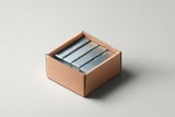 Open cardboard box containing four neatly stacked bundles of metal staples on a neutral surface.