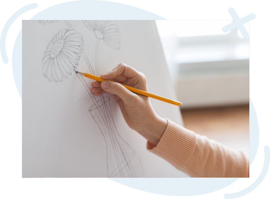 Close-up of a hand sketching flowers in a vase with a yellow pencil on white paper.