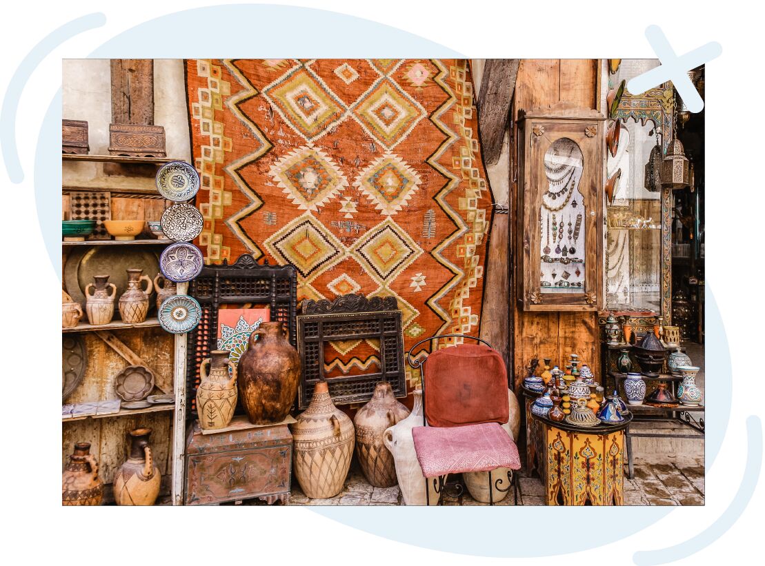 Colorful market stall displaying Moroccan-style crafts: a large orange geometric rug hangs as a backdrop, surrounded by patterned ceramic plates, clay jugs, carved wooden frames, a glass cabinet with jewelry, painted tagine pots, and an ornate side table 