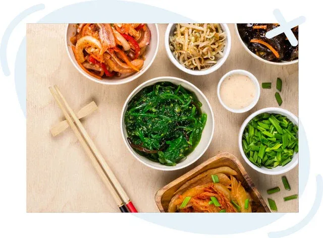 Assorted small bowls of Korean-style side dishes on a light wooden table with chopsticks, including seaweed salad, kimchi, bean sprouts, sliced scallions, and dipping sauce.