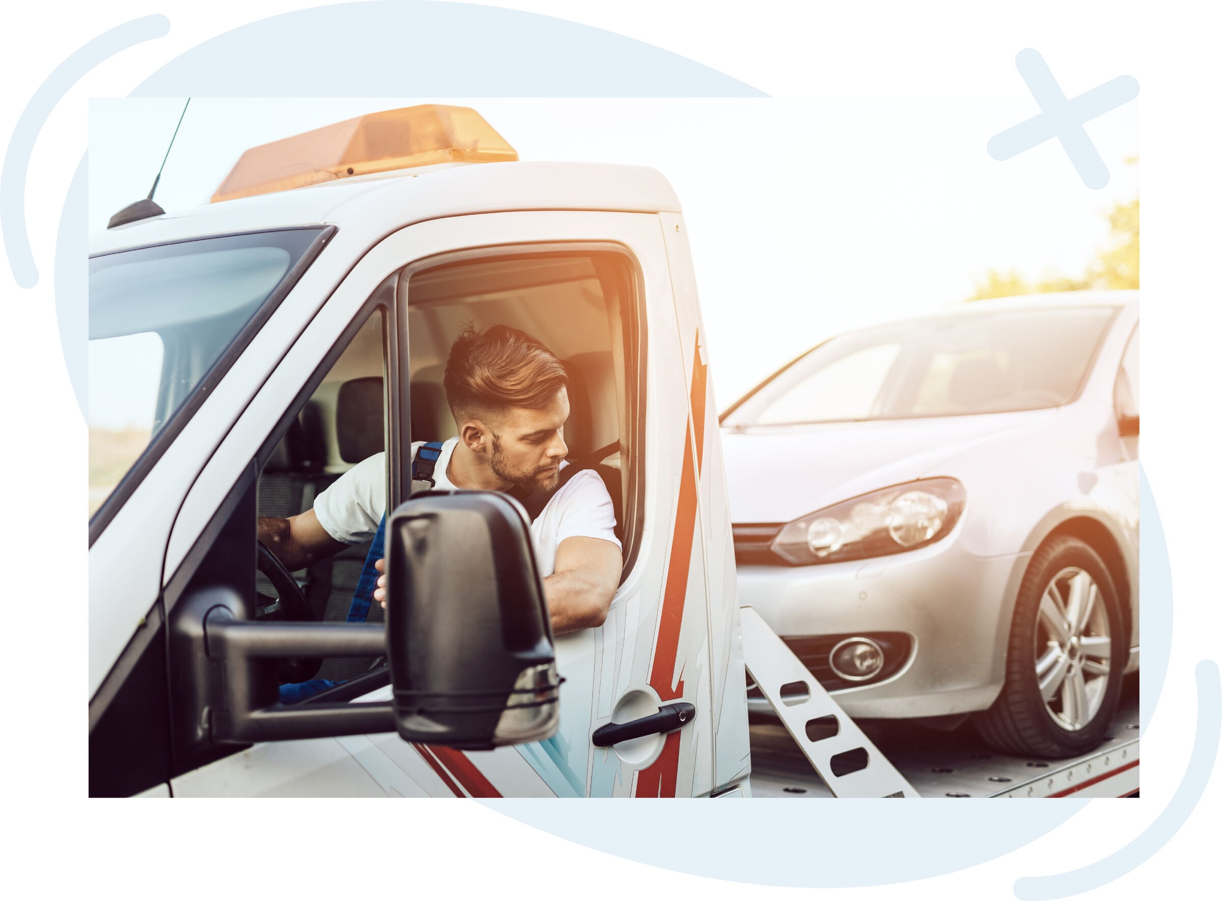 Tow truck driver looking back while loading a silver car onto a flatbed at sunset.