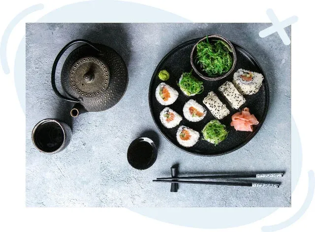 Top-down view of a Japanese meal with assorted sushi rolls on a black plate, seaweed salad, pickled ginger, wasabi, chopsticks, and a cast-iron teapot with teacups on a gray stone surface.